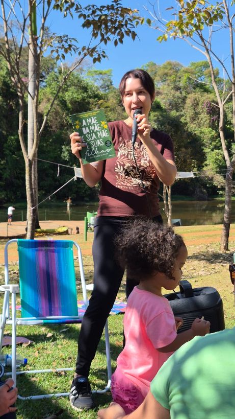Mediadora lê ao microfone segurando o livro, com uma criança em primeiro plano